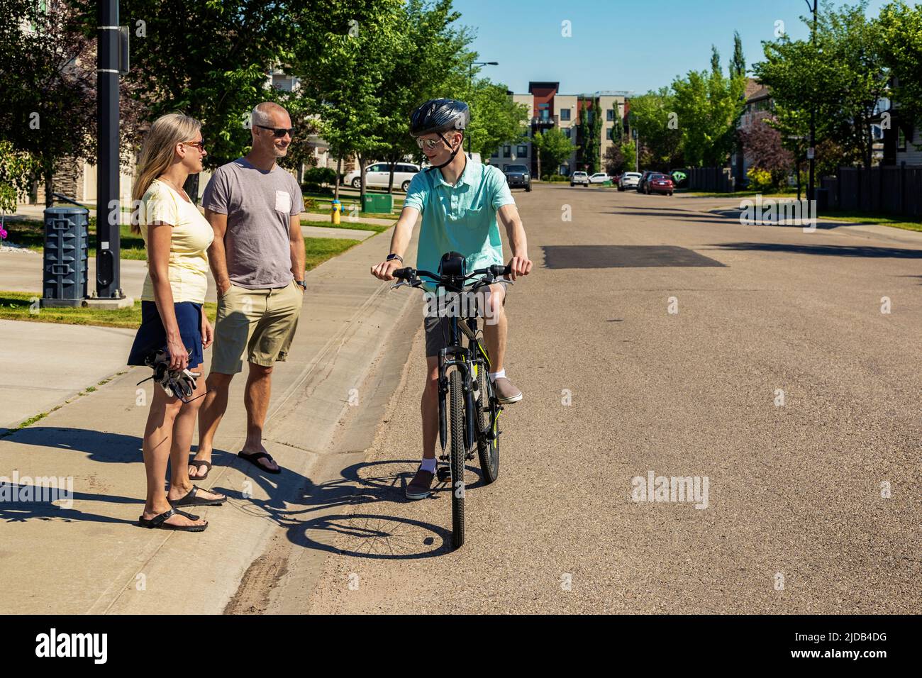 Joven listo para montar su bicicleta por una calle residencial, parando a hablar con sus padres que están de pie en la acera Foto de stock