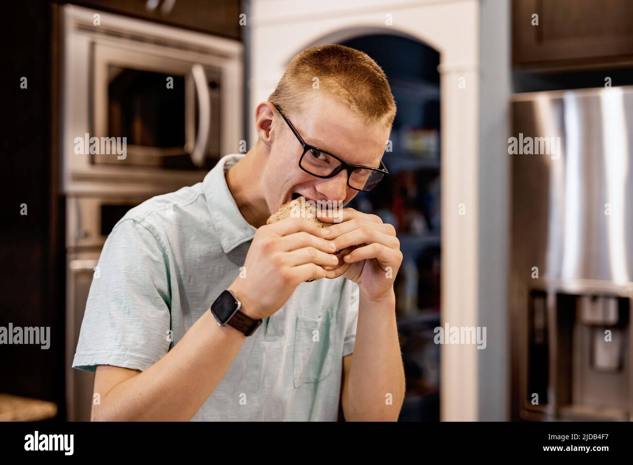 Joven de pie en la cocina en casa y comiendo un sándwich, mirando a la cámara; Edmonton, Alberta, Canadá Foto de stock