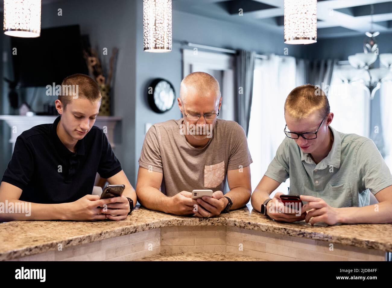 Padre y dos hijos se sientan en la isla de la cocina en casa usando sus teléfonos inteligentes; Edmonton, Alberta, Canadá Foto de stock