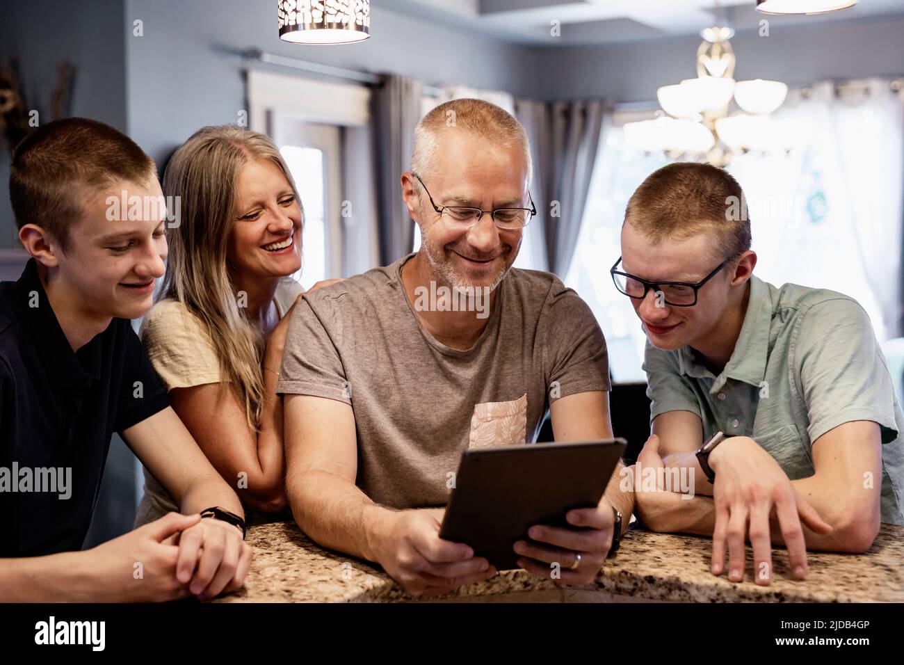 Los padres y dos hijos se sientan en la isla de la cocina en casa usando una tableta y viendo contenido juntos; Edmonton, Alberta, Canadá Foto de stock