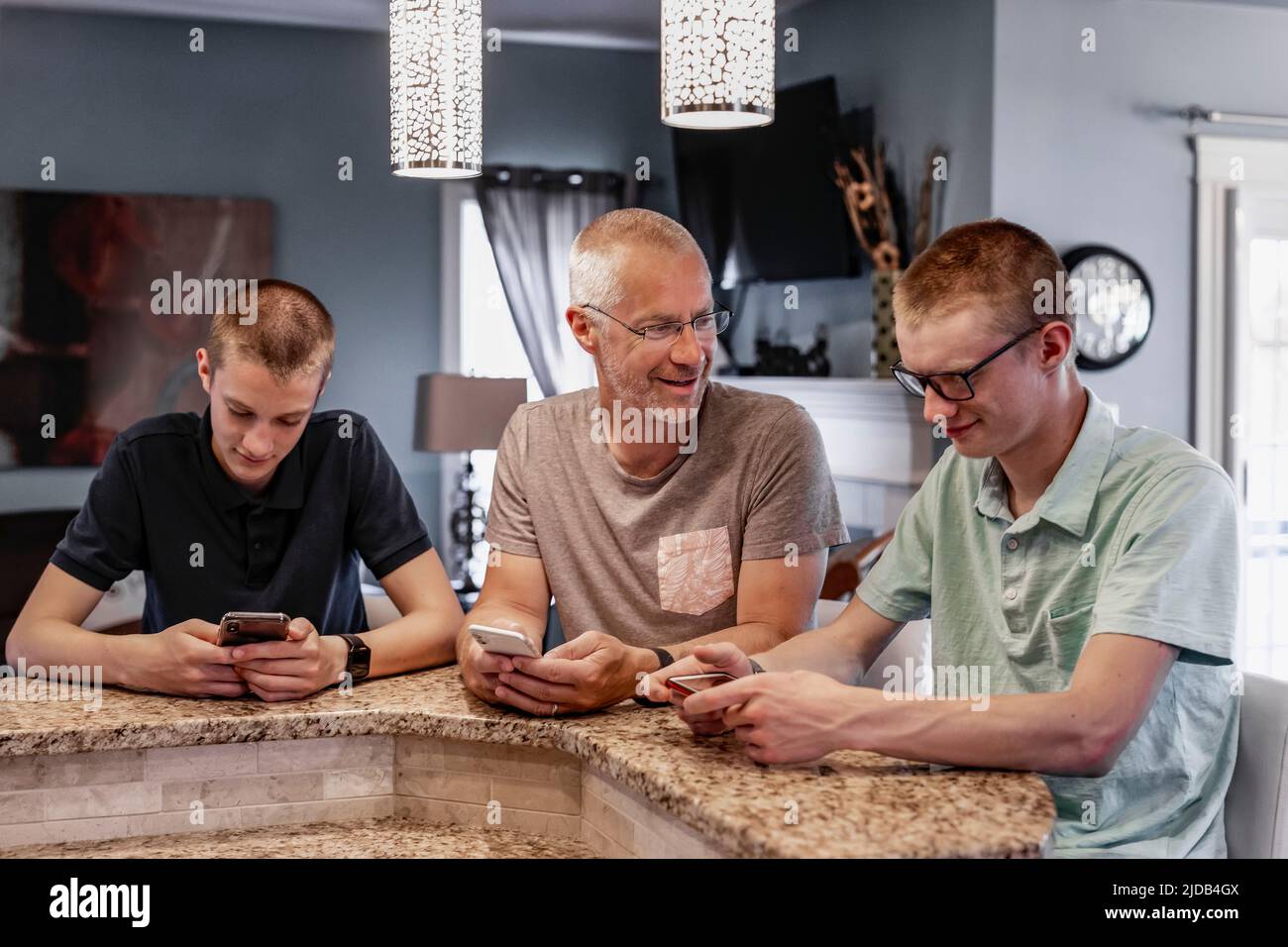 Padre y dos hijos se sientan en la isla de la cocina en casa usando sus teléfonos inteligentes, el padre tratando de hablar con ellos; Edmonton, Alberta, Canadá Foto de stock