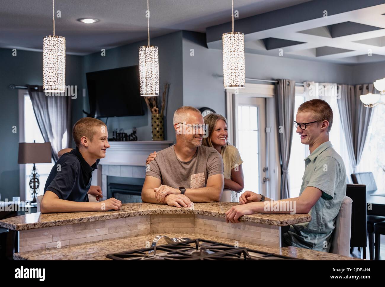 Padre y madre con dos hijos se sientan en la isla de la cocina en casa hablando juntos; Edmonton, Alberta, Canadá Foto de stock