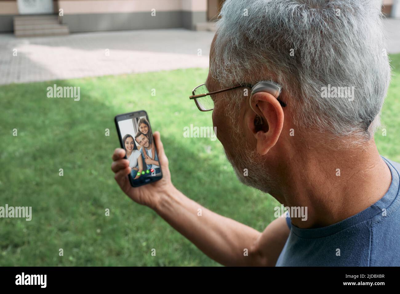 Un hombre mayor con un audífono detrás del oído se comunica con su familia a través de una comunicación de vídeo a través de un smartphone. Vida humana completa con audífonos Foto de stock