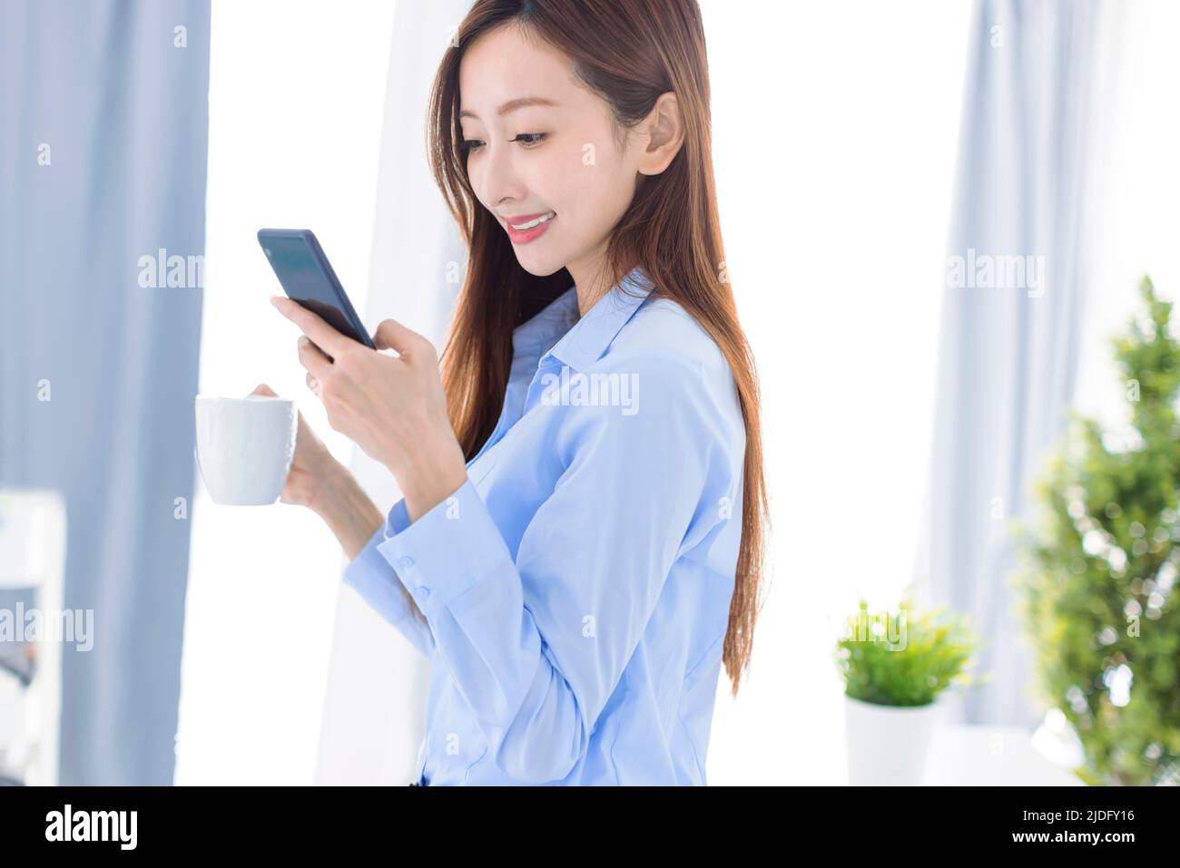 Joven mujer de negocios tomando café y viendo el teléfono inteligente en la oficina Foto de stock