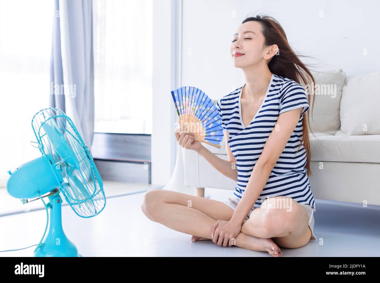 Mujer joven Relájese en la onda de la sala de estar con el ventilador de mano y disfrutando del flujo de aire del ventilador . Concepto de calor de verano Foto de stock