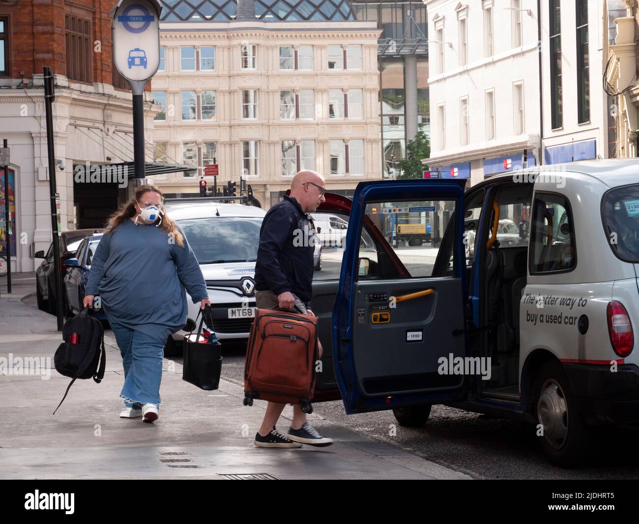 El taxista negro de Londres ayuda a cargar el equipaje en la cabina de espera, con una mujer que lleva una máscara de covid del PPE Foto de stock