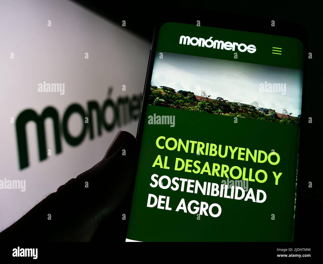 Persona que sostiene el celular con la página web de la empresa Monomeros Colombo Venezolanos S.A. en la pantalla delante del logo. Enfoque en el centro de la pantalla del teléfono. Foto de stock