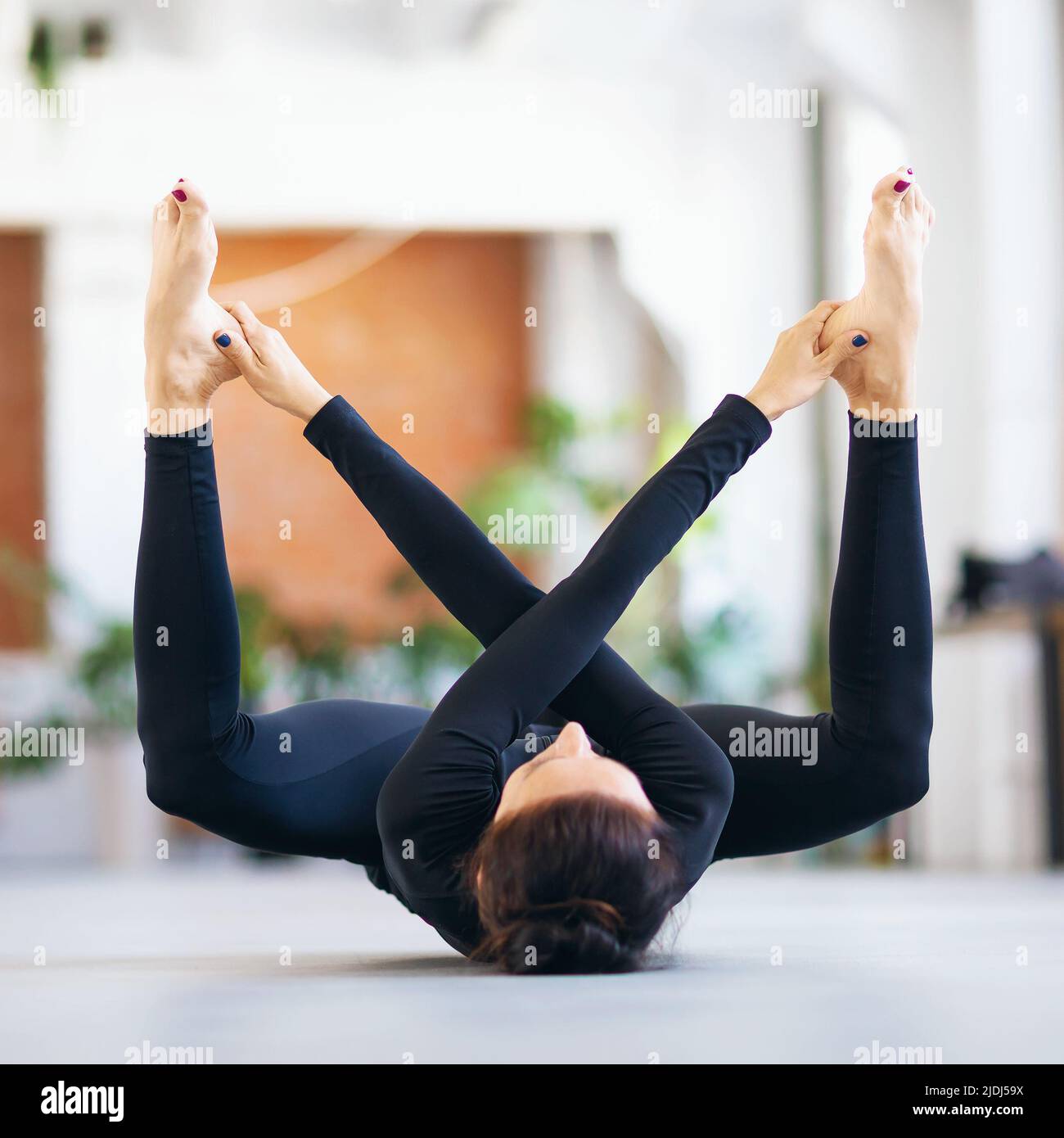 Mujer en ropa deportiva negra practicando yoga haciendo ejercicio supta rudrasana, haciendo ejercicio mientras se encuentra tumbado en una colchoneta en la habitación Foto de stock