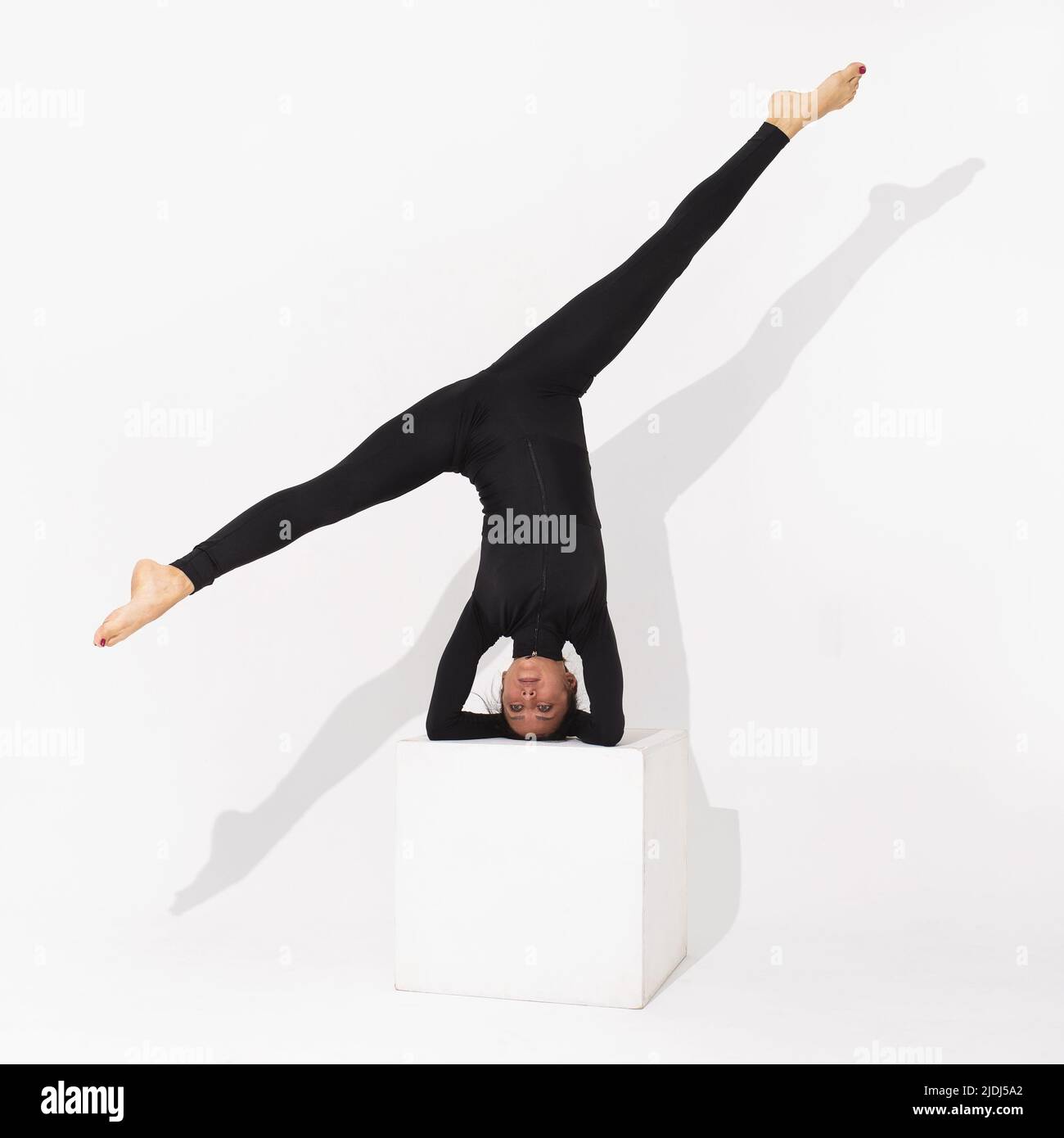 Una mujer en ropa deportiva negra practicando yoga realiza el ejercicio Salamba Shirshasana, la postura asana invertida o el cabecero, se coloca sobre un cubo en una b blanca Foto de stock