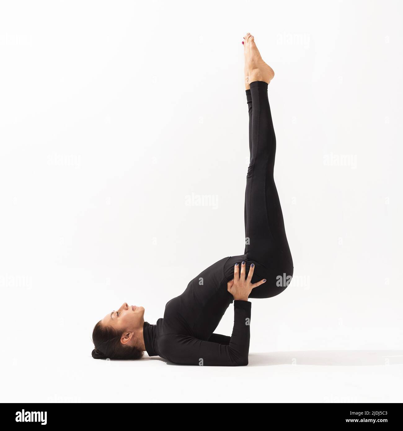 Una mujer de ropa deportiva negra que practica yoga realiza el ejercicio Viparita Karani, puesta de vela invertida, sobre un fondo blanco Foto de stock