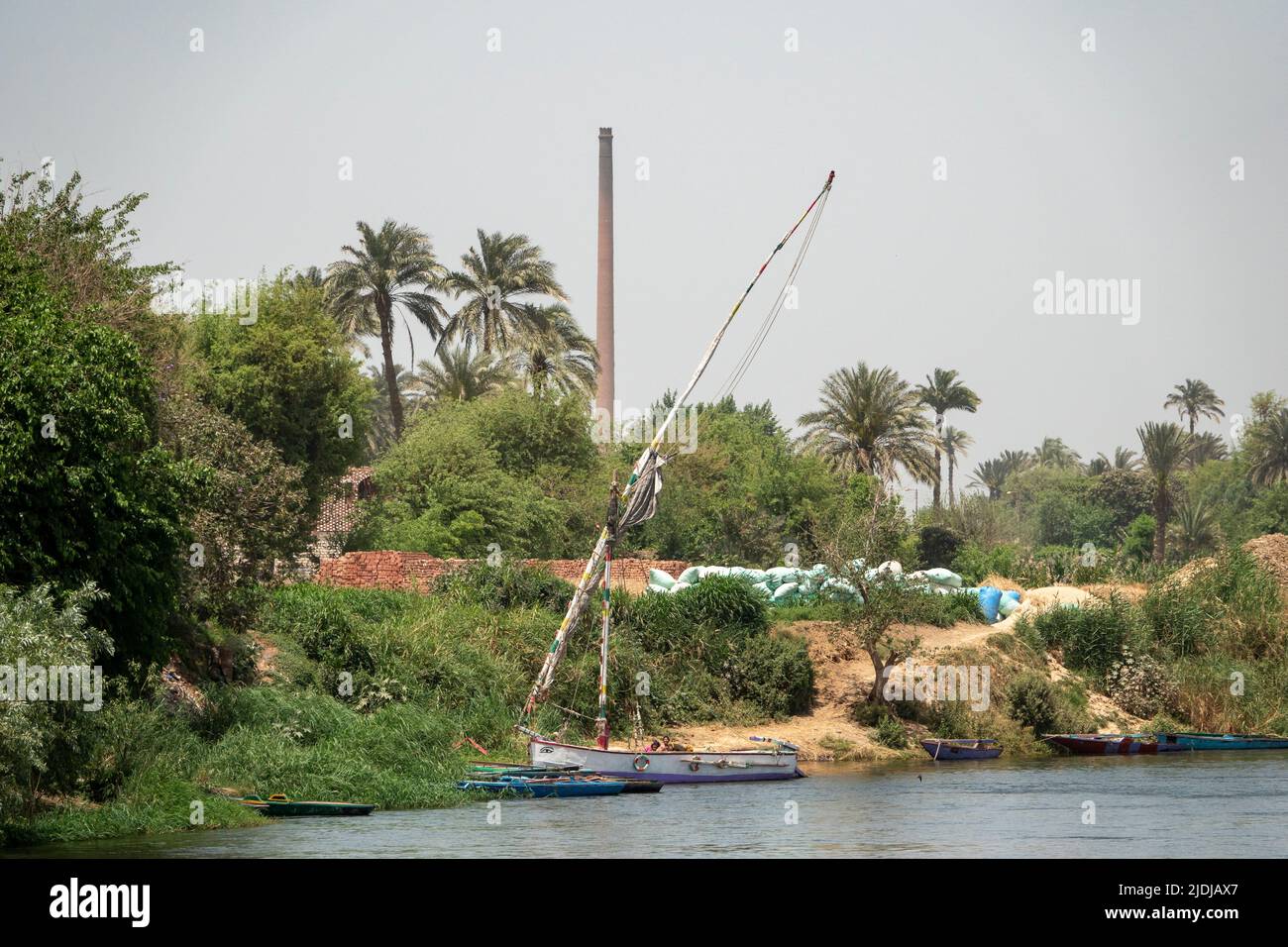 Una felucca tradicional con varios pequeños barcos amarrados en las cañas a orillas del río Nilo con ladrillo y chimenea en el fondo Foto de stock