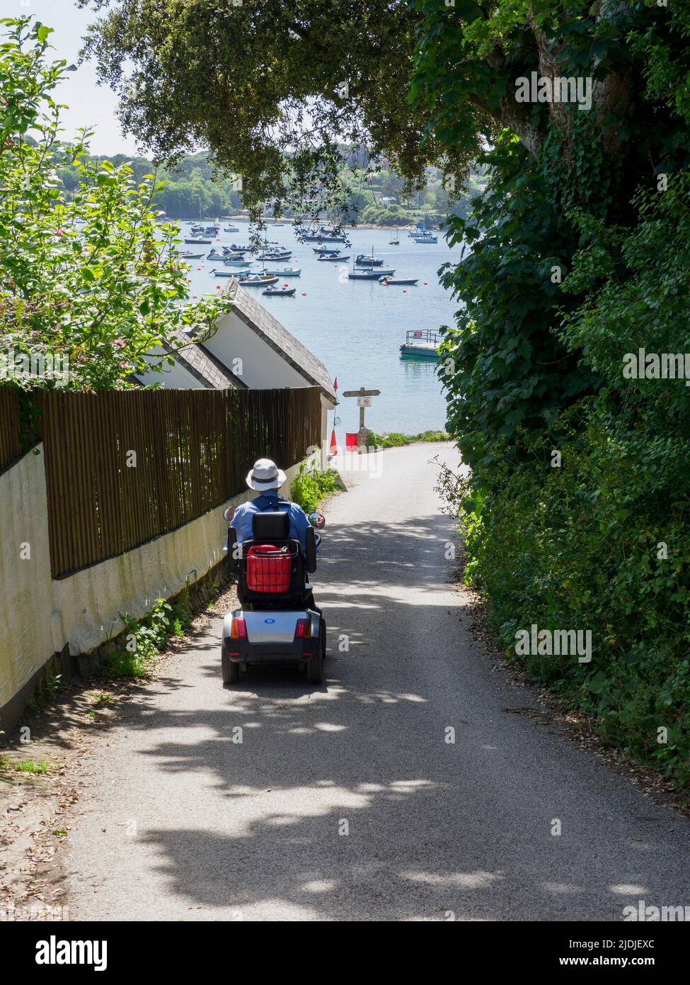 Ciudadano mayor que monta en una scooter de movilidad por la carretera a la playa, Helford Passage, Cornwall, Reino Unido Foto de stock