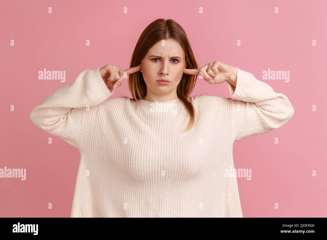 No quiero oír esto. Retrato de una mujer rubia infeliz de pie y poniendo el dedo en sus orejas, mirando la cámara, usando un suéter blanco. Estudio en interior grabado aislado sobre fondo rosa. Foto de stock