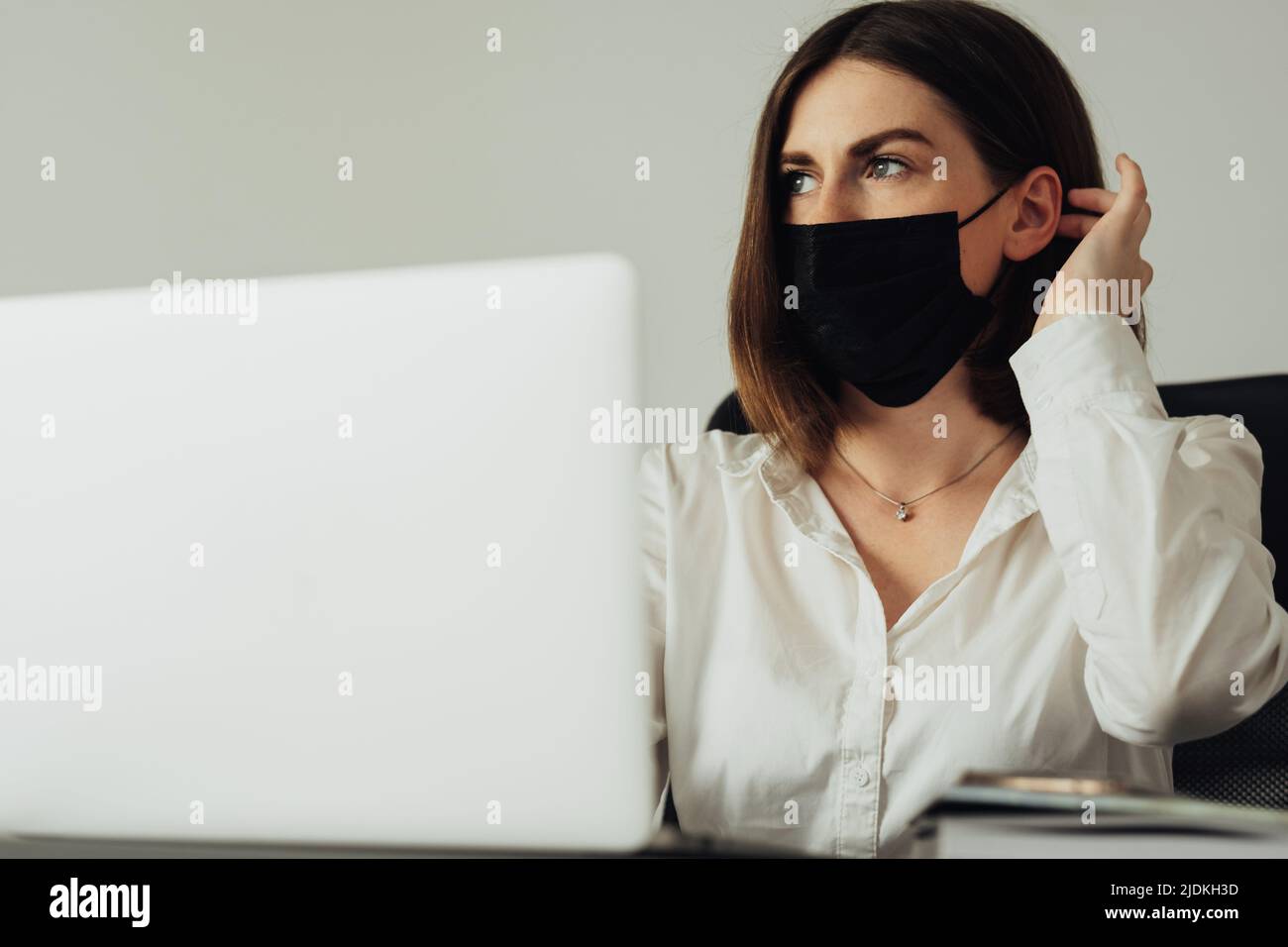 Retrato de una empleada sentada en la oficina, una mujer joven con máscara de protección y trabajando en el ordenador portátil Foto de stock