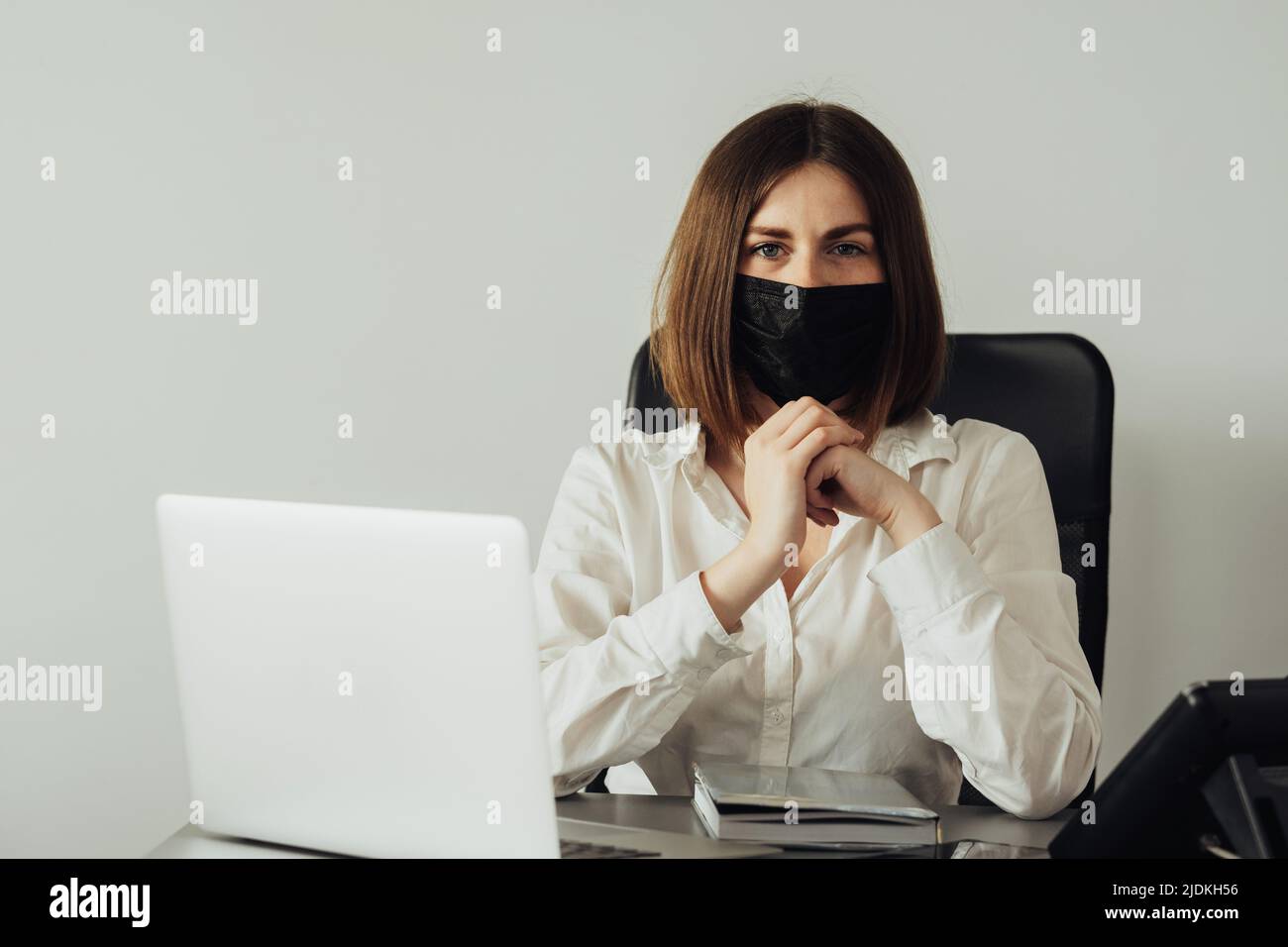 Retrato de la joven directora de oficina mirando hacia la cámara, mujer confiada en la máscara de protección sentada en el lugar de trabajo Foto de stock