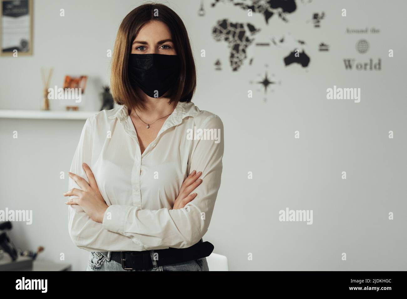Retrato de la joven mujer caucásica con máscara protectora de pie en la oficina con brazos de sujeción, espacio de copia Foto de stock