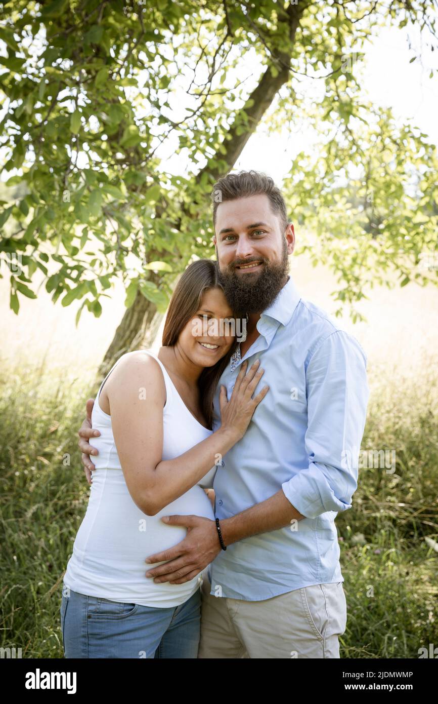 una mujer embarazada joven y con camisa blanca está de con su novio con barba y camisa azul en un de flores altas y acurrucado Fotografía de stock -