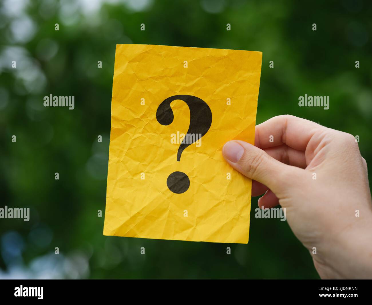 Una mujer sosteniendo un papel de nota amarillo con un signo de interrogación en su mano sobre un fondo de follaje. Primer plano. Foto de stock