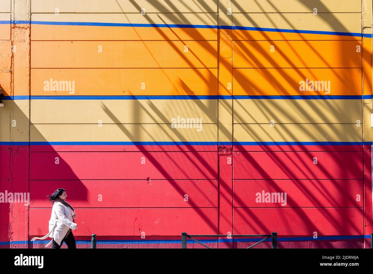 Señora con un abrigo blanco caminando frente a una colorida pared. Sombra de una estructura metálica. Bruselas. Foto de stock