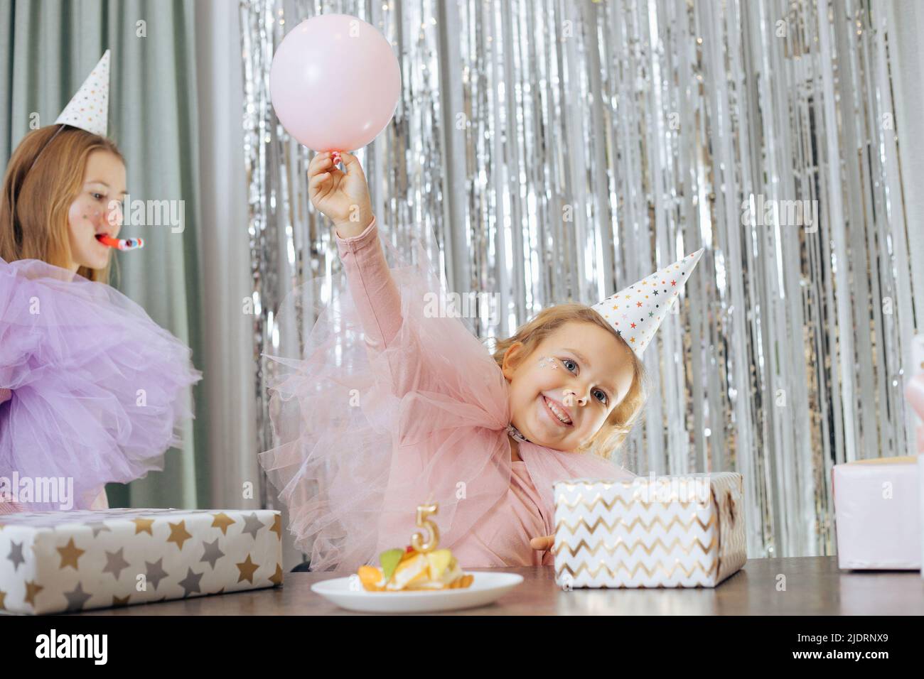 Hermoso cumpleaños. Muy alegre bebé de 5 años en sombrero de fiesta levantó su mano con globo delante de ella es pastel de cumpleaños y muchas cajas con regalos Foto de stock