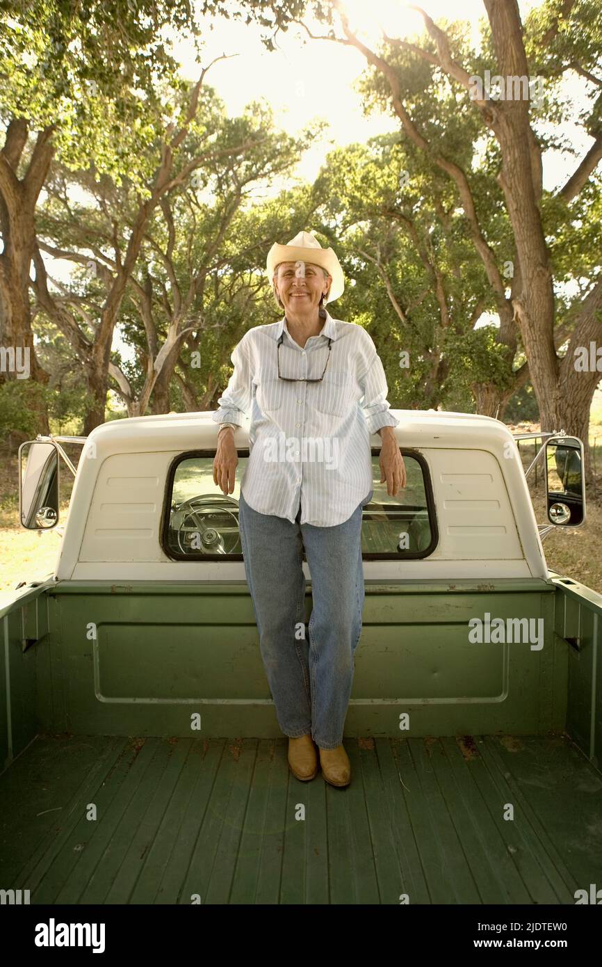 Mujer (de 45 a 65 años) con sombrero vaquero apoyado en la cabina de una camioneta vintage aparcada en la carretera del rancho bordeada de la madera de algodón de Fremont envejecida (Populus Foto de stock