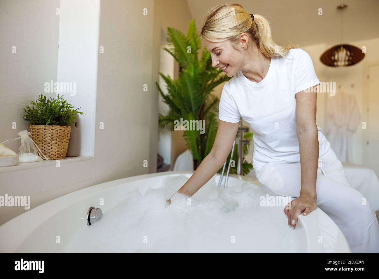 Mujer rubia bastante joven haciendo baño caliente con espuma en casa. Tiempo de relajación. Acogedor cuarto de baño. Foto de stock