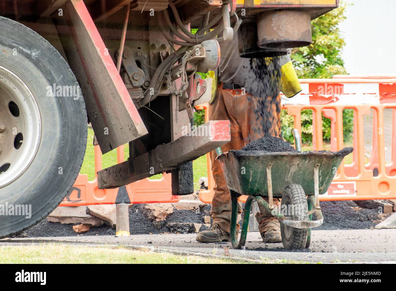 Un obrero espera junto a una carretilla mientras se llena de asfalto fresco de un camión. El asfalto es para reparaciones a una calle que ha sido excavada Foto de stock