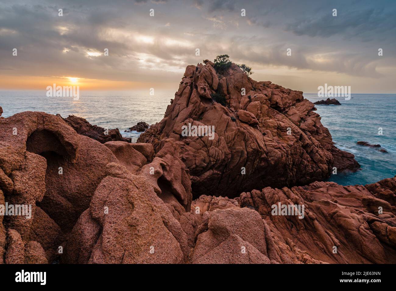Salida del sol con el espectacular cielo en la bahía de Cocorocci con las rocas de pórfido rojo, guijarros grises y Monte Cartucceddu en la costa este de Cerdeña Foto de stock