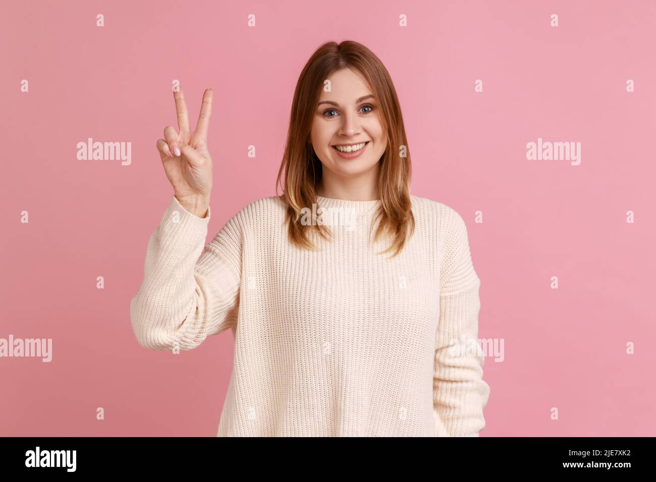 Mujer mostrando victoria o gesto de paz, sonriendo ampliamente, regocijándose afortunado ganando, sintiéndose optimista sobre el éxito futuro, usando suéter blanco. Estudio de interior grabado aislado sobre fondo rosa. Foto de stock