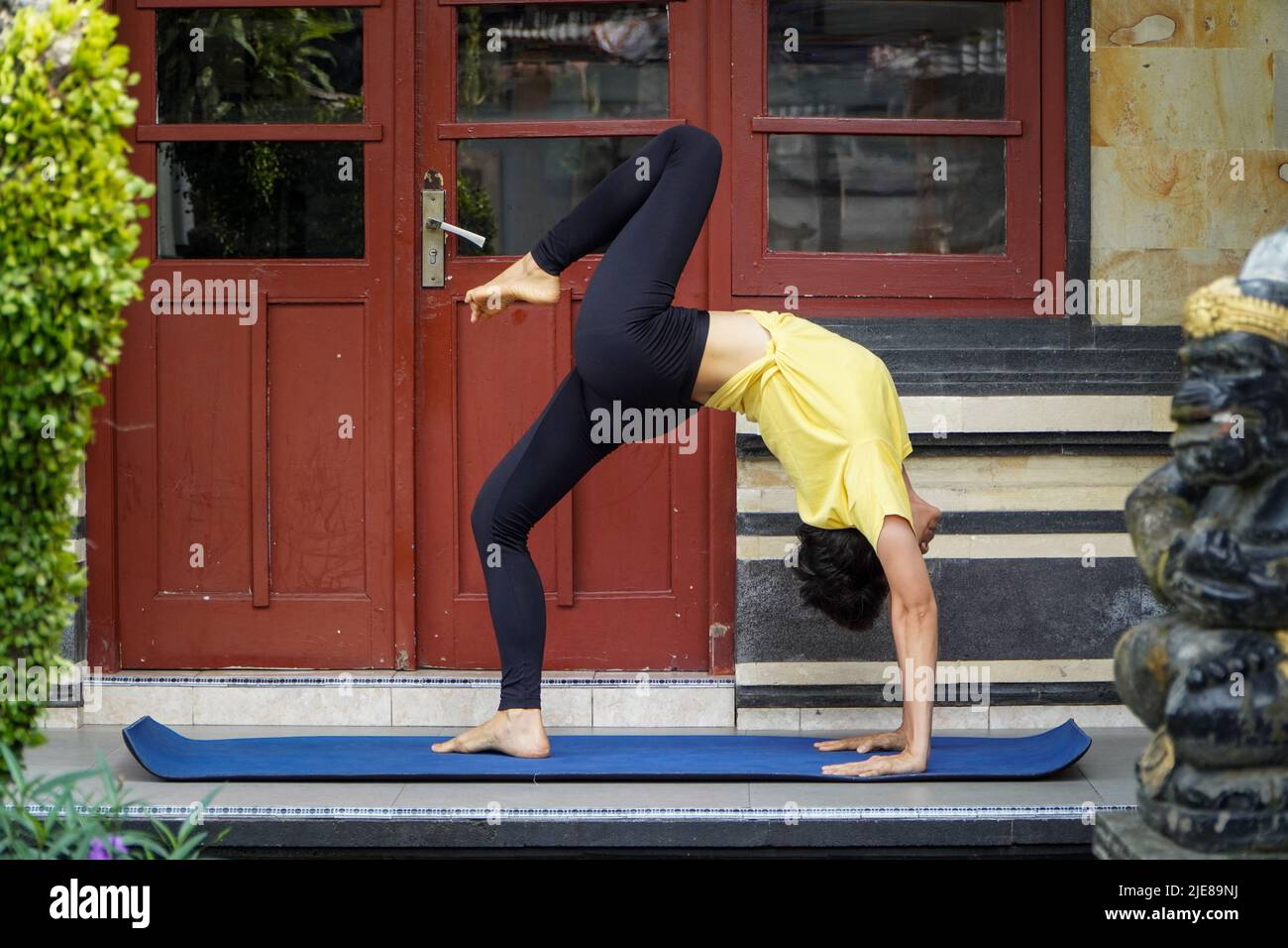 En su terraza, una joven asiática con un aspecto impresionante está practicando Yoga mientras se divierte un corte de pelo corto, una camisa amarilla y leggings negros. S Foto de stock