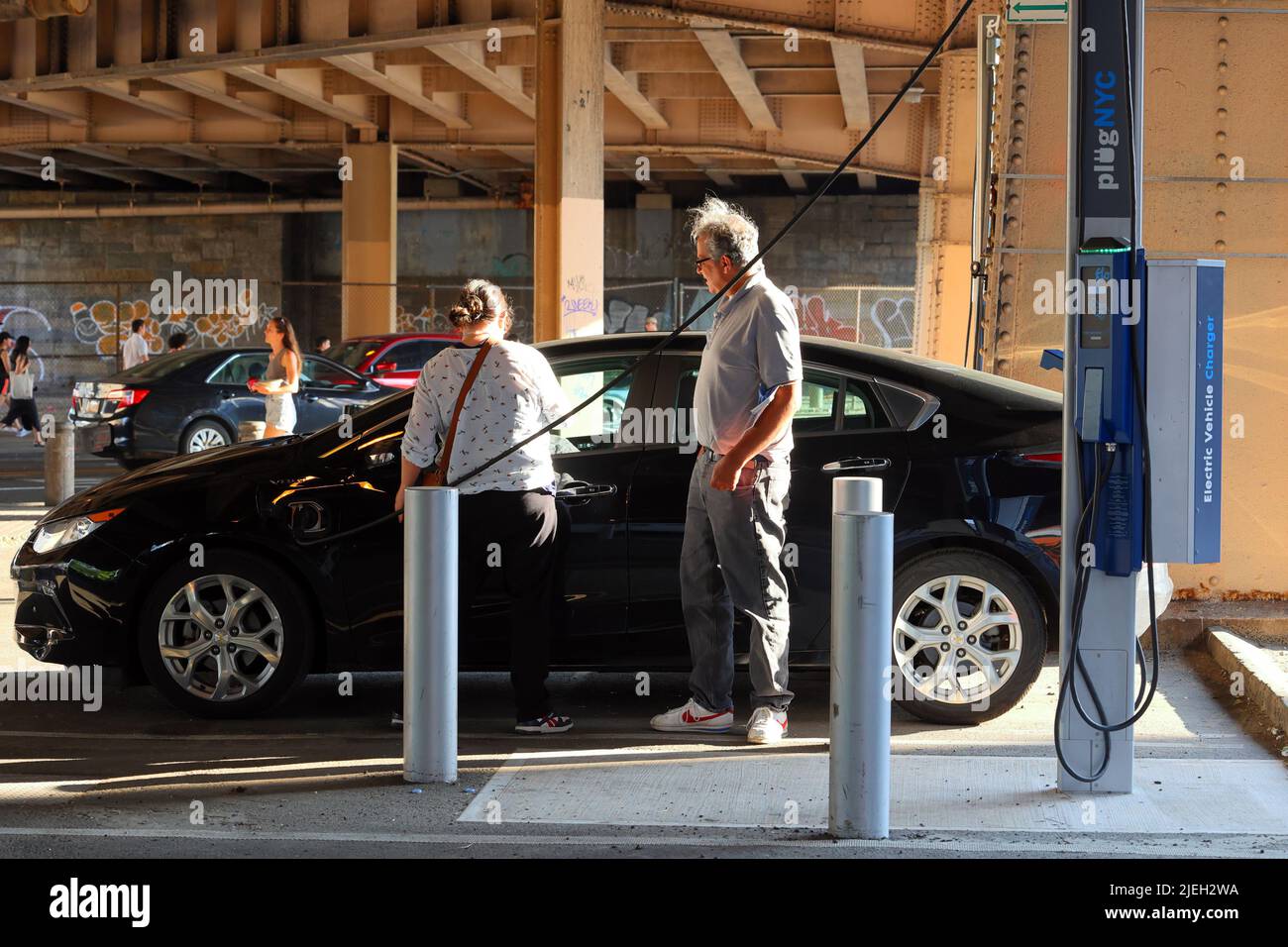 Personas que utilizan un raro cargador de vehículo eléctrico PlugNYC curbside para recargar su coche eléctrico en la red pública de New York City.The es operado por Flo Foto de stock