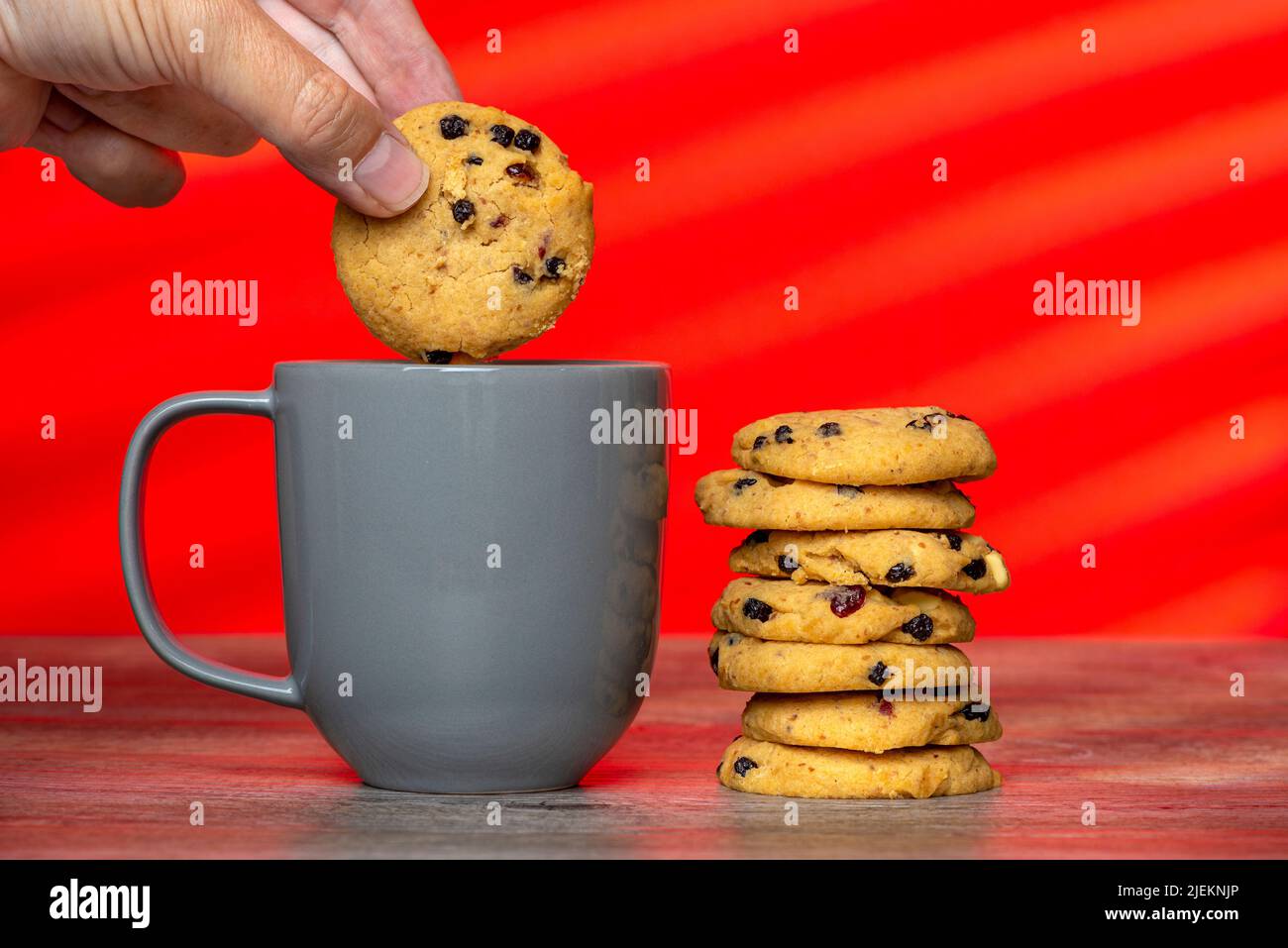 pila de galletas caseras y una taza gris sobre un fondo rojo con una mano dunking una galleta. Foto de stock