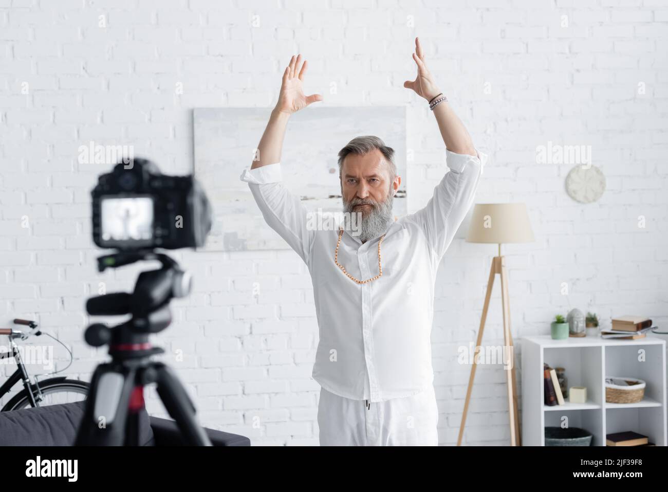 mentor espiritual barbudo meditando con las manos levantadas cerca de una cámara digital borrosa en casa Foto de stock