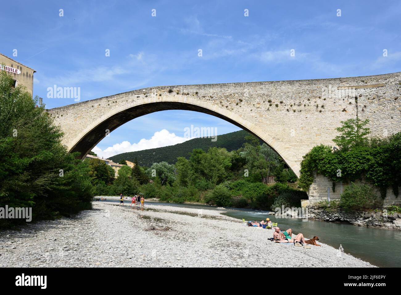 Los turistas tomando el sol y relajándose a lo largo del río Aigues bajo el Puente Medieval Single Span, conocido como el Puente Romano, Nyons Drôme Provence Francia Foto de stock