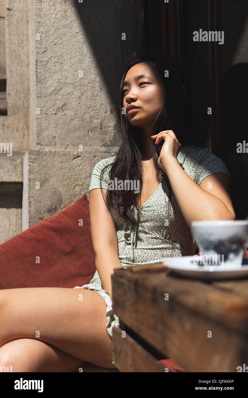 Mujer asiática de ensueño sentada mirando a la mesa con una taza de café en la terraza del café al aire libre el soleado día de verano y disfrutando el fin de semana Foto de stock