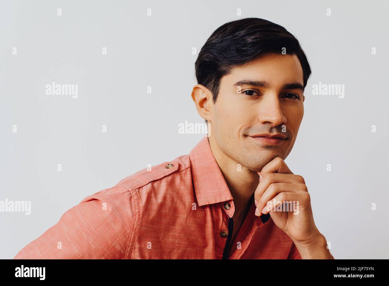 Headshot pensativo hombre joven adulto latino con la mano en la barbilla pelo negro y camisa rosa sobre fondo gris mirando la cámara de estudio de disparo Foto de stock