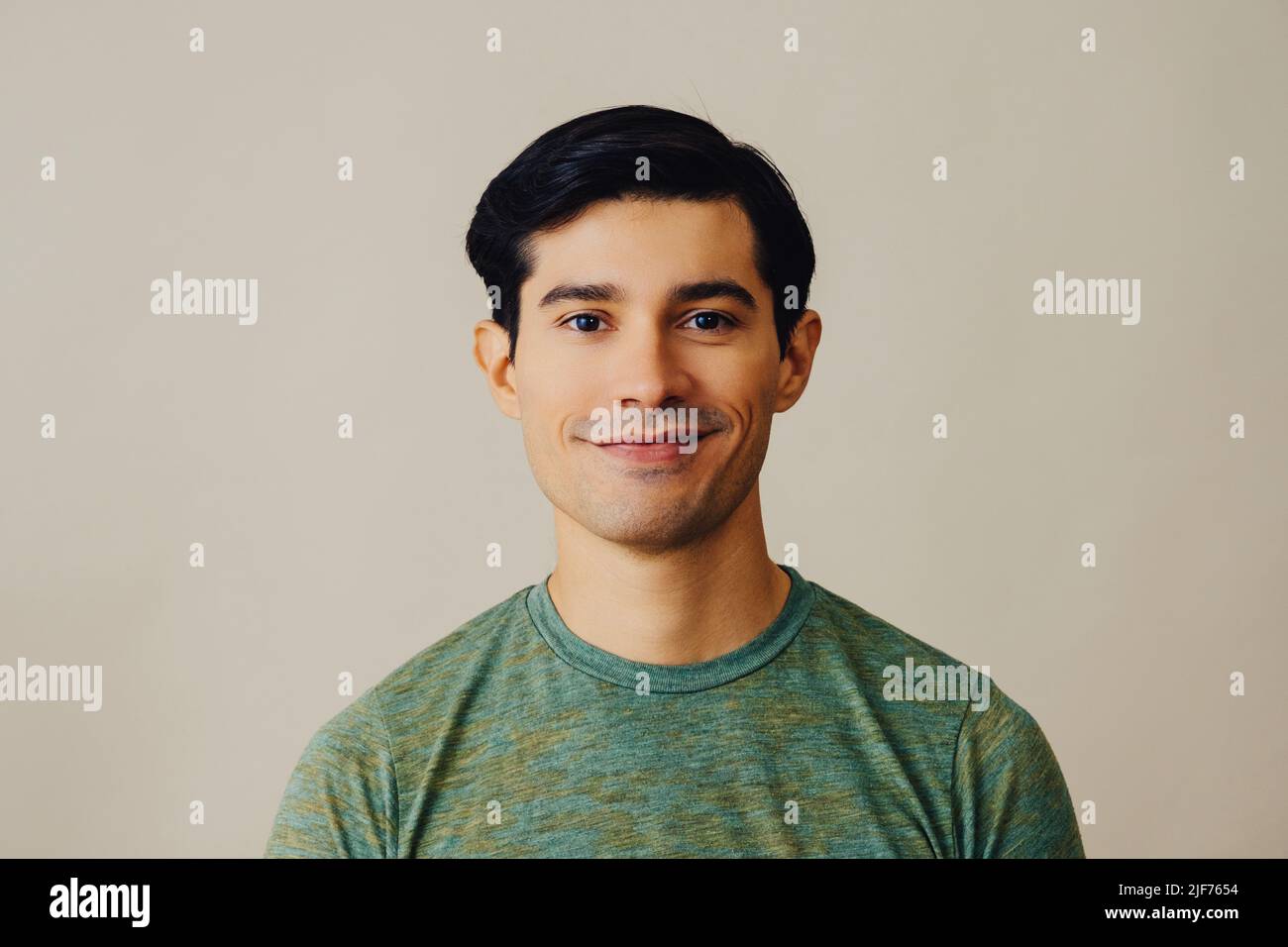 Headshot hombre latino pelo negro sonriente guapo adulto joven camiseta verde sobre fondo gris mirando la cámara de estudio de disparo Foto de stock