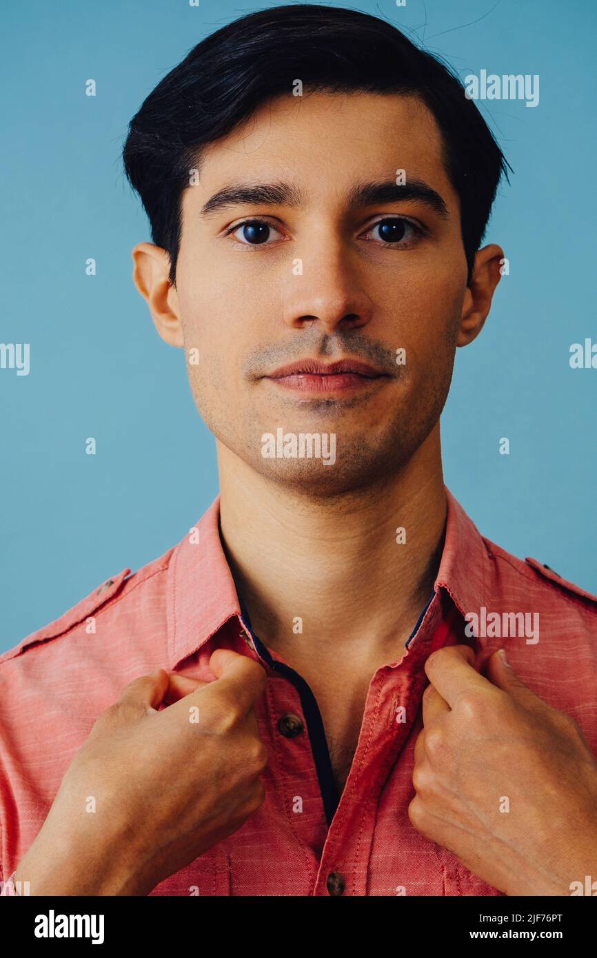 Headshot hombre hispano latino pelo negro sonriente hermoso joven adulto con camisa rosa sobre fondo azul mirando la cámara de estudio de disparo Foto de stock