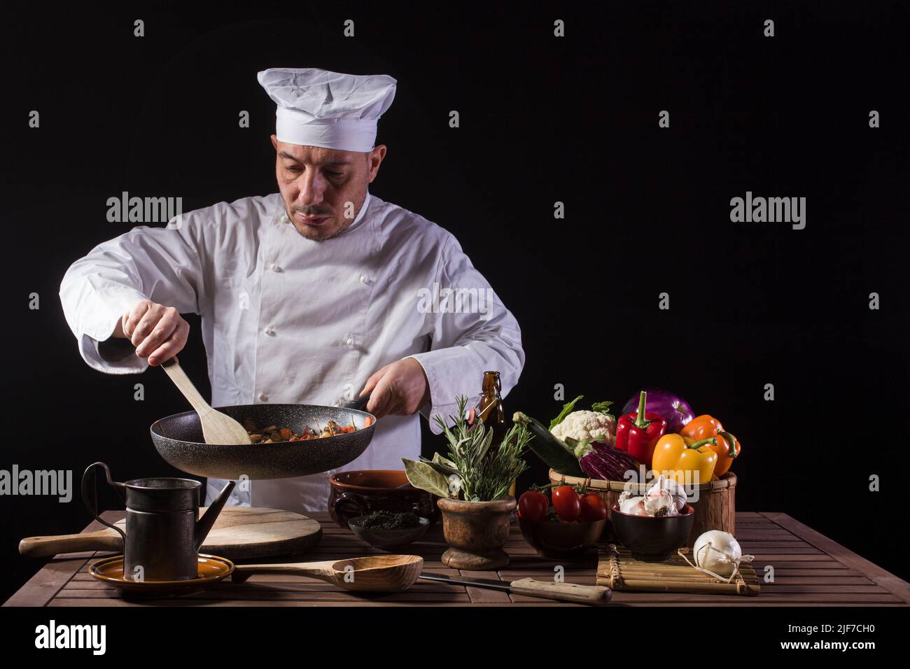 Chef con uniforme blanco y sombrero con cuchara mezcla los ingredientes en la sartén antes de servir mientras trabaja en la cocina de un restaurante Foto de stock