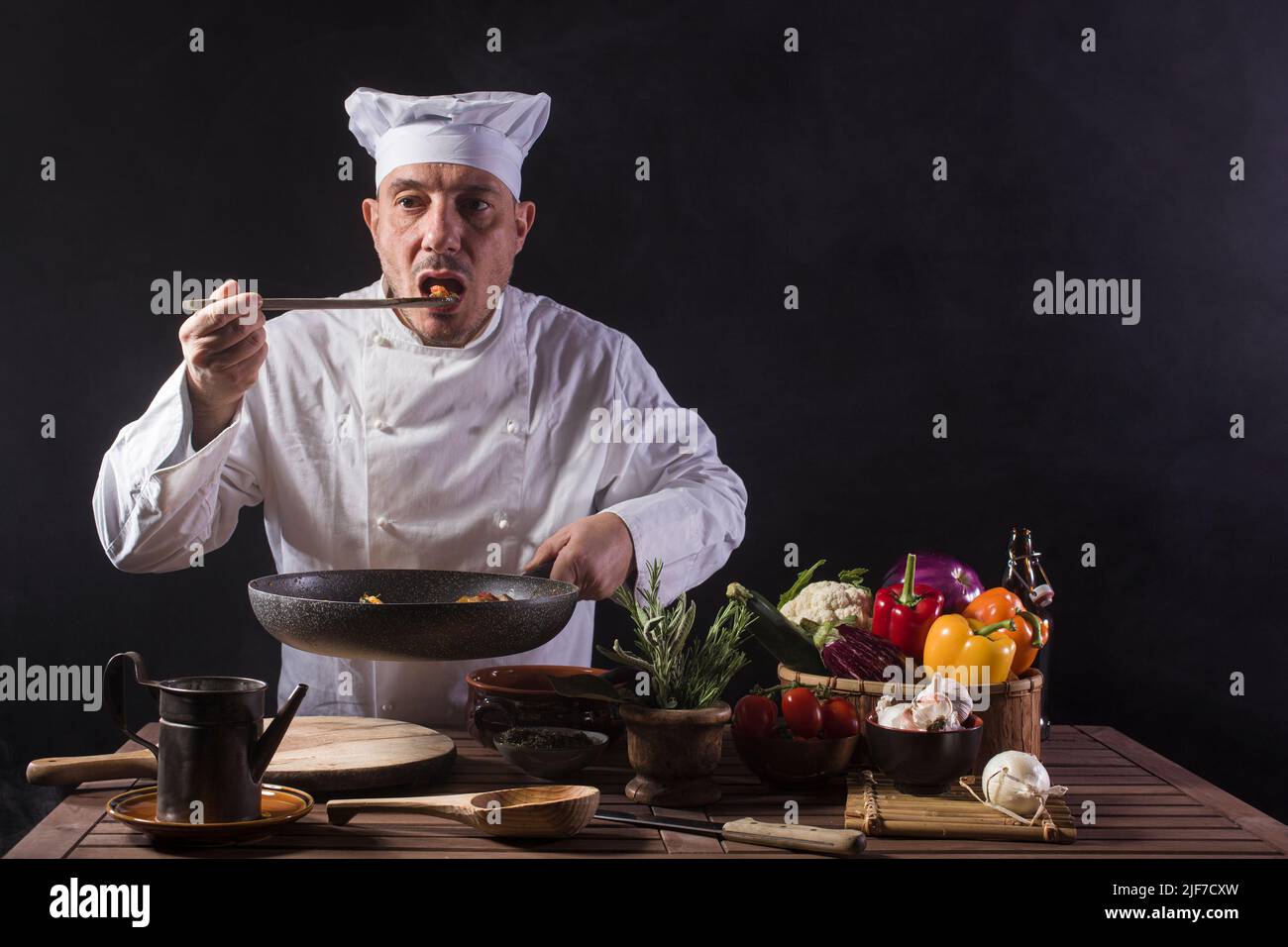 Chef masculino con uniforme blanco es probar la comida utilizando un ladle de madera en la cocina de un restaurante antes de servir mientras trabaja en una cocina de restaurante Foto de stock