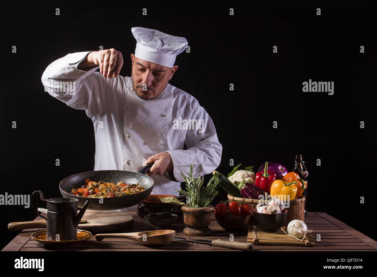 Chef masculino con uniforme blanco y sombrero poniendo sal y hierbas en el plato de comida con verduras antes de servir mientras trabaja en la cocina de un restaurante Foto de stock