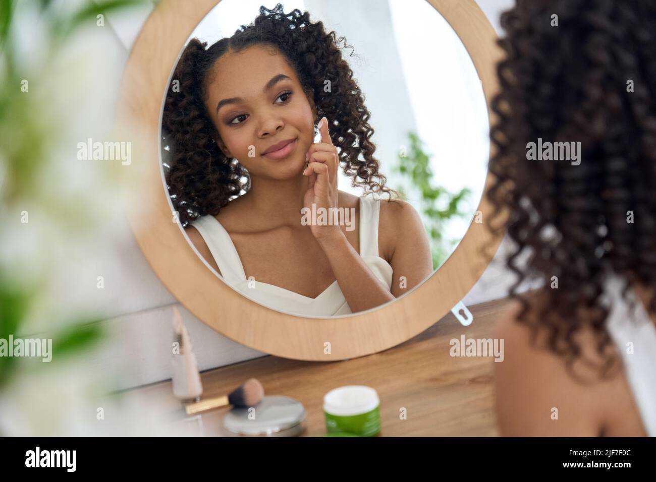 Chica africana adolescente mirando en el espejo del baño aplicando crema facial en la cara. Foto de stock
