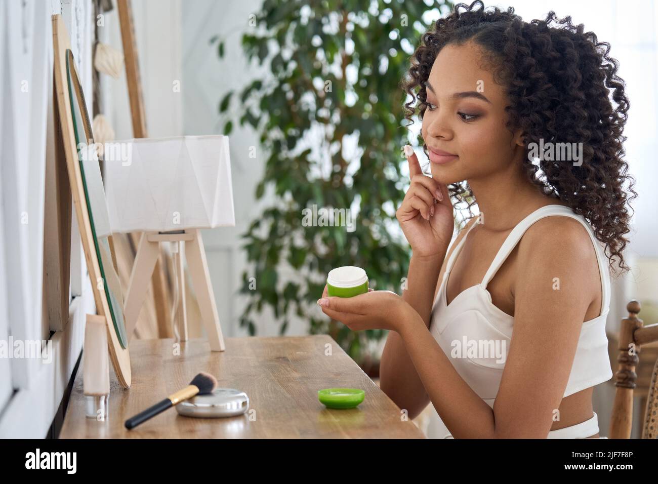 Chica afroamericana adolescente sentada mirando el espejo aplicando crema facial. Foto de stock