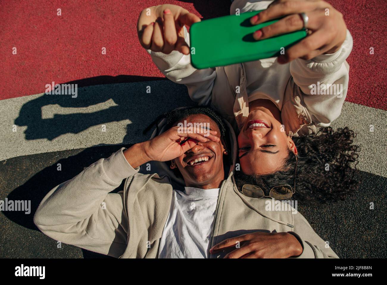 Amigos jóvenes y felices tomando selfie a través del teléfono inteligente mientras están tumbados en el patio de recreo Foto de stock