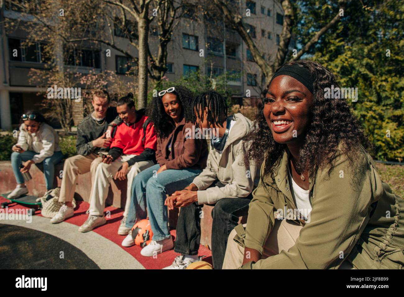 Retrato de una sonriente joven sentada con amigos multirraciales en una pared de retención en el patio de recreo Foto de stock