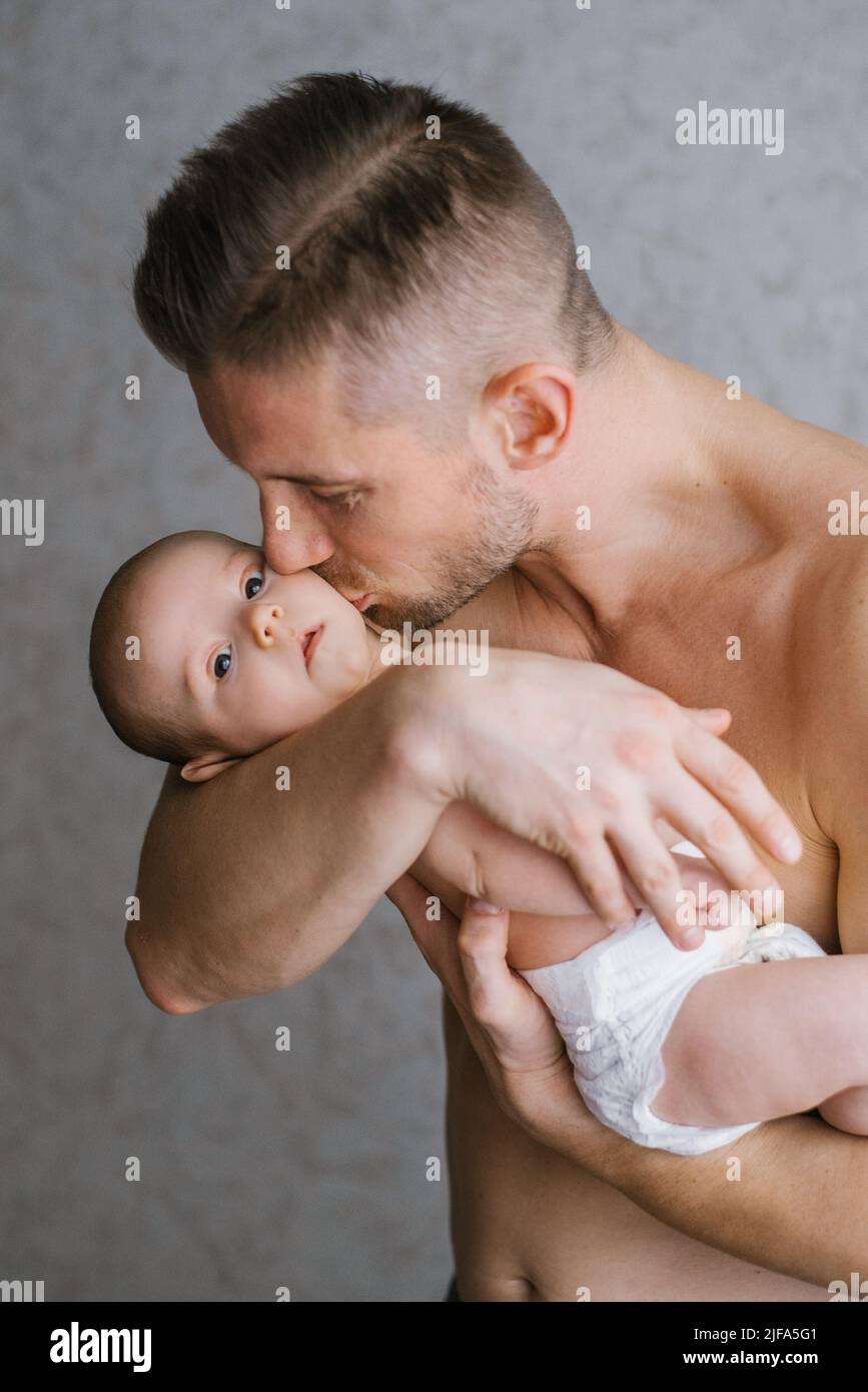 Papá sostiene a su bebé en sus brazos en casa y besa al bebé en la mejilla Foto de stock