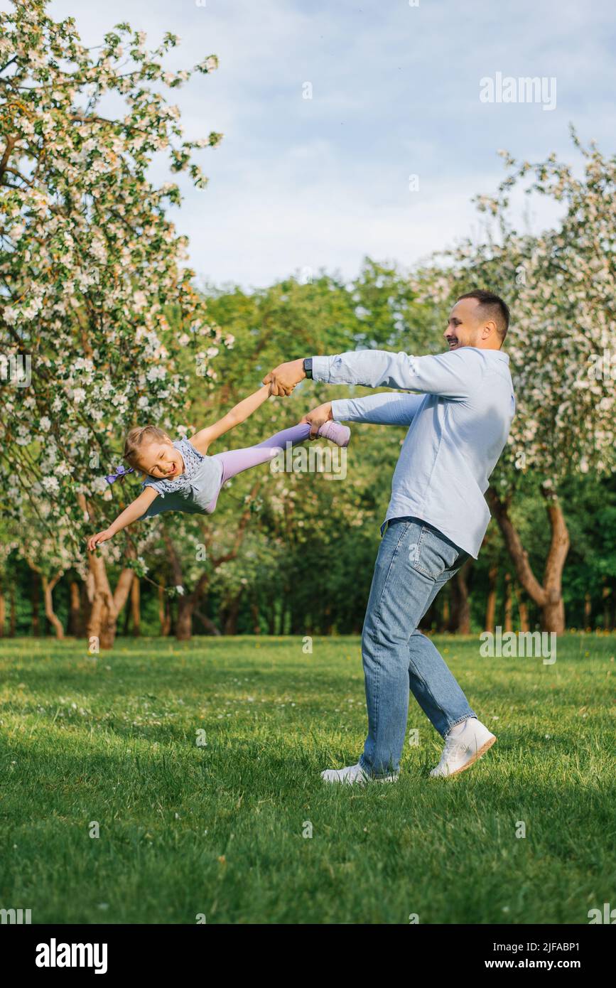 Alegre padre tuerce a su hija sonriente con la mano y con la pierna a su alrededor durante un paseo por el parque en primavera Foto de stock