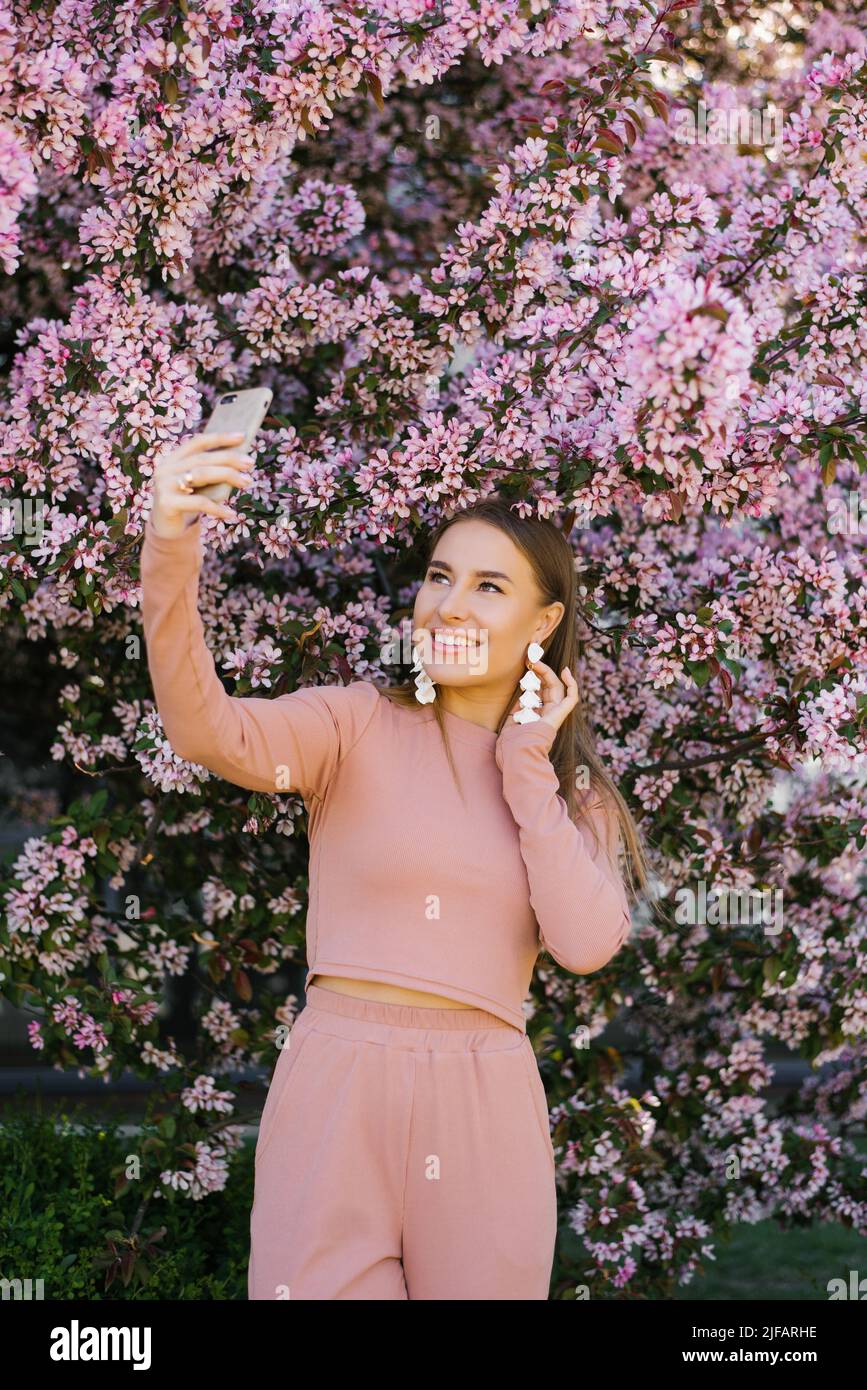 Una mujer joven toma un selfie en su teléfono rodeado de flores de manzana rosa. Temporada de primavera Foto de stock