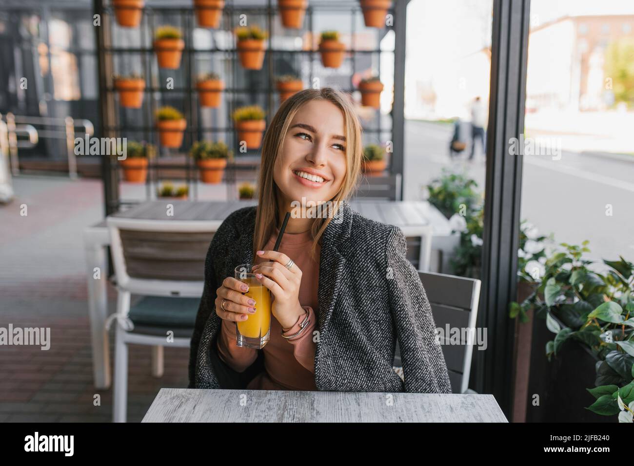 Una joven feliz está sentada en una mesa en un café de verano y sosteniendo un vaso de jugo de mango en su mano Foto de stock