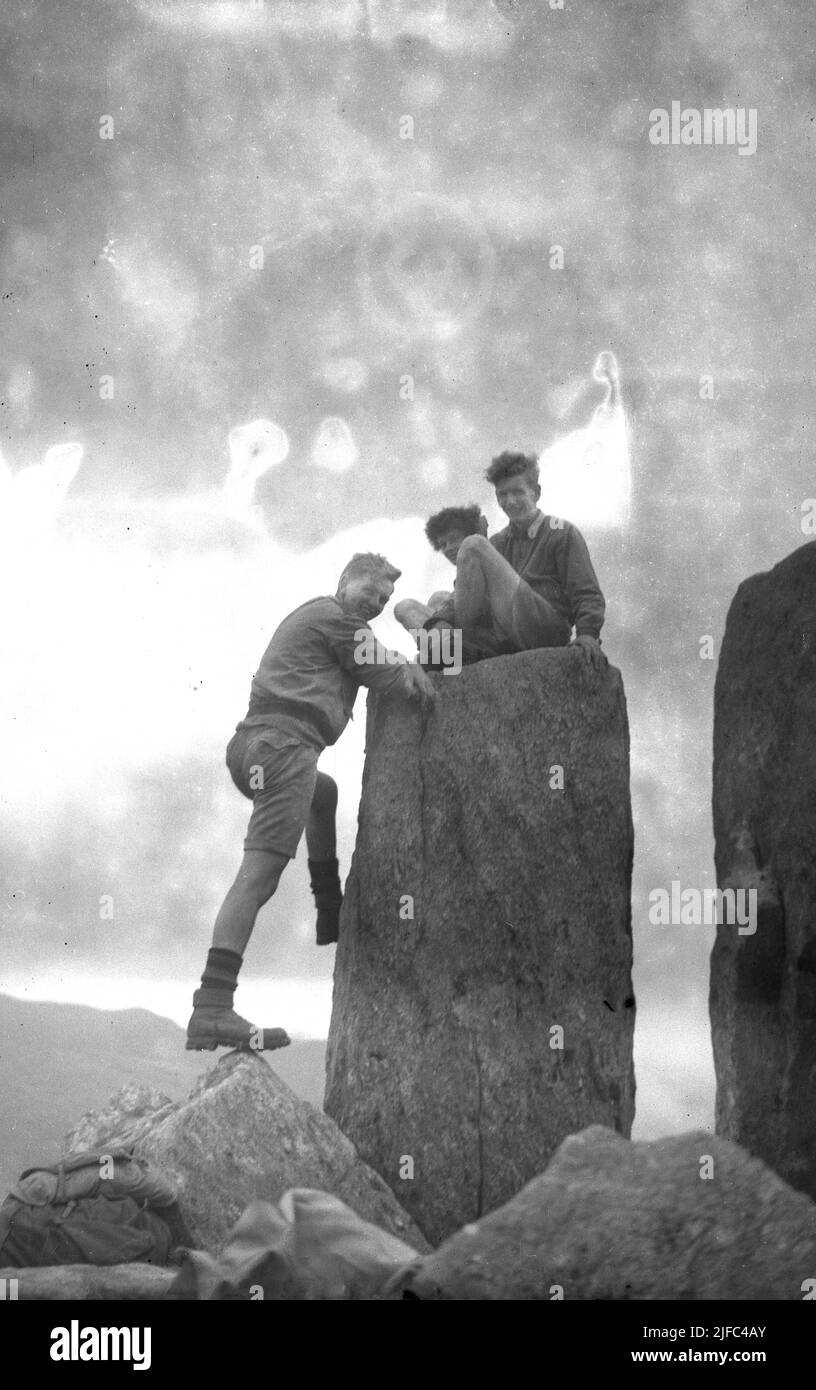 1952, histórico, tres hombres caminantes en la cima de la montaña de Tryfan, Snowdonia, Gales, con dos sentados en la parte superior de uno de los monolitos gemelos de Adán y Eva (Sion a Sian en Wlesh) un par de rocas de tres metros de altura. Foto de stock
